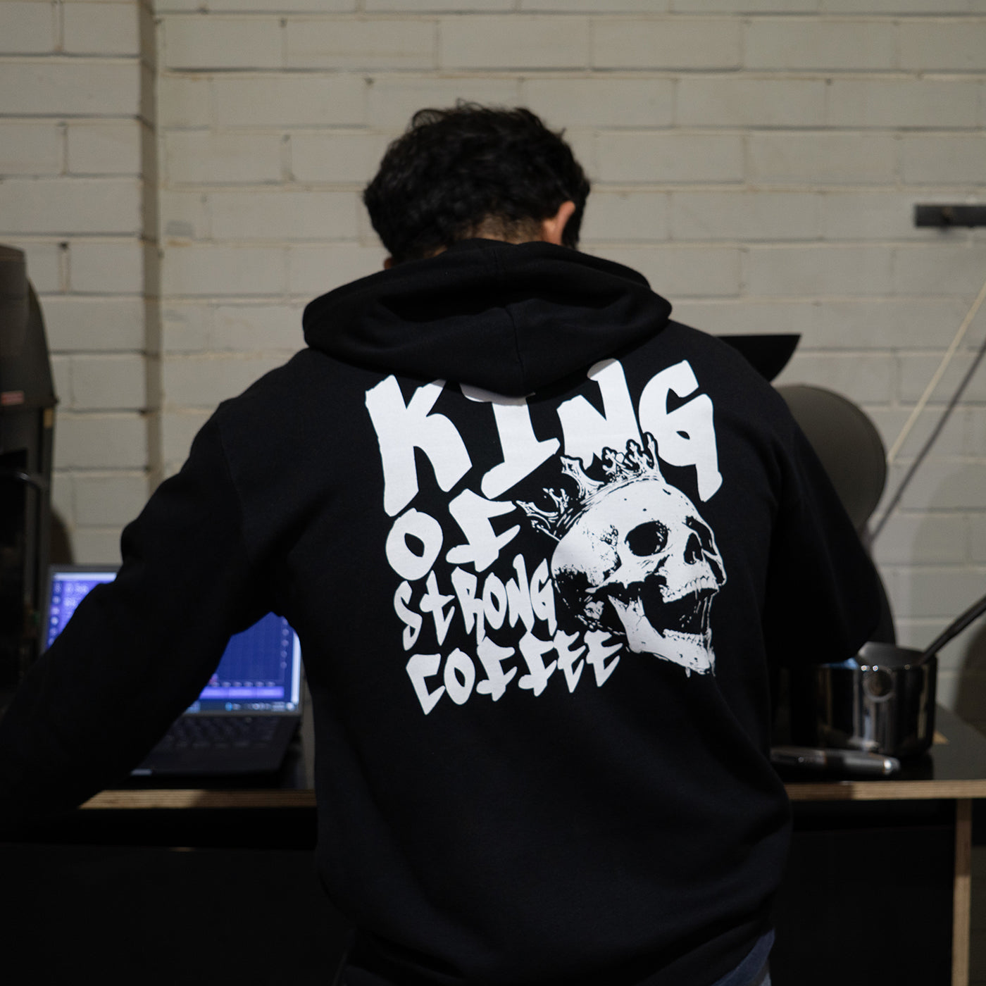 Person wearing a black hoodie with white text and skull design, sitting in a room with a laptop and coffee mug.